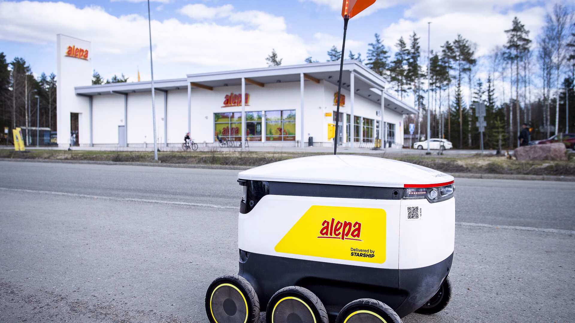 A Starship robot with a yellow Alepa sticker crossing a road